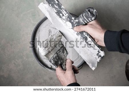 Top view of hands transferring wall filler from a putty knife onto a wide taping knife above a bucket. Concept of drywall finishing, wall repair and DIY home renovation work.
