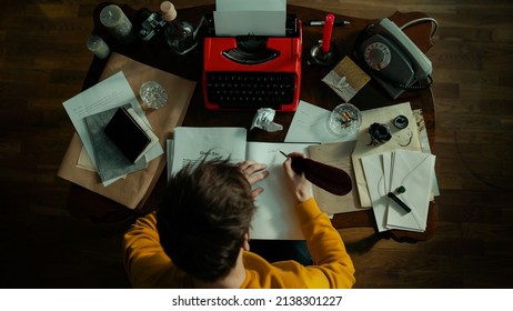 Top View Of A Handsome Brunette Male Writer Working On His Next Chapter Behind An Antique Table With Vintage Objects Covering It Like A Red Typewriter, Telephone, Notebooks, An Ashtray, Ink And Quill.