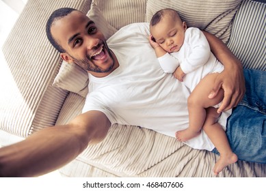 Top view of handsome young Afro American father smiling while making selfie with his little baby sleeping in his arms, lying on sofa at home