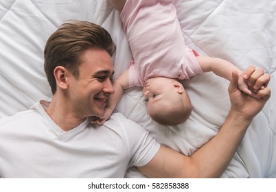 Top View Of Handsome Young Dad And His Cute Baby Looking At Each Other While Lying On Bed