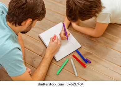 Top view of handsome young father and his cute little son drawing while lying on a wooden floor in the room - Powered by Shutterstock