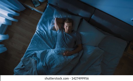 Top View of Handsome Young Man Sleeping Cozily on a Bed in His Bedroom at Night. Blue Nightly Colors with Cold Weak Lamppost Light Shining Through the Window. - Powered by Shutterstock