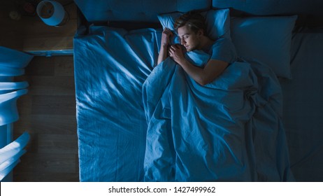 Top View Of Handsome Young Man Sleeping Cozily On A Bed In His Bedroom At Night. Blue Nightly Colors With Cold Weak Lamppost Light Shining Through The Window.