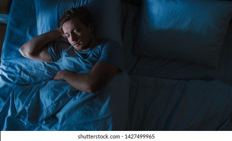 Top View of Handsome Young Man Sleeping Cozily on a Bed in His Bedroom at Night. Blue Nightly Colors with Cold Weak Lamppost Light Shining Through the Window.