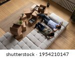 Top view happy couple tenants homeowners relaxing on couch on moving day in living room with cardboard boxes, man and woman resting after relocation in first own apartment, mortgage or rent