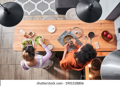 Top View Of The Happy Curly Multiracial Woman Grated Cheese At The Pasta While Preparing Dinner. Her Bestie Pouring Salad At The Table. Young Women Friends Cooking Meal Together At Home. Stock Photo