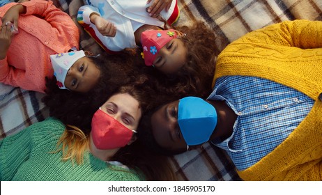 Top View Of Happy Diverse Family In Safety Mask Relaxing On Blanket During Picnic. Mixed Race Parents And Little Children Wearing Protective Mask Lying On Blanket Outdoors