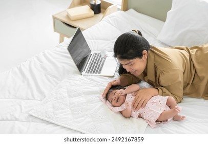 Top view of happy family asian mother and newborn baby lying and playing together on white bed. Mother working on laptop and nursing her infant girl or boy at home. Care and working concept. - Powered by Shutterstock