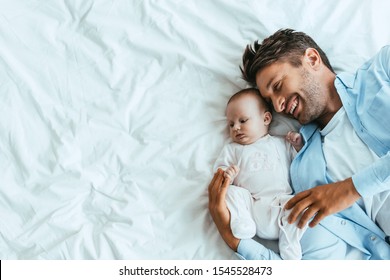 Top View Of Happy Father Hugging Adorable Baby While Lying On White Bedding