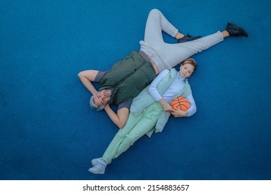 Top View Of Happy Father And Teen Daughter Lying And Looking At Camera Outside At Basketball Court.