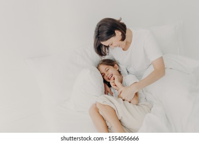 Top View Of Happy Funny Girl In Pyjamas And Her Caring Mom Pose On Bed, Play Together In Bed, Enjoy Softness And Comfort, Express Positive Emotions. Family, Morning Time And Relaxation Concept