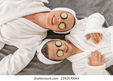 Top view of happy mother and daughter in bathrobes lying with cucumber slices on eyes, mom and cute preteen female child making beauty treatments together at home, relaxing on bed after spa day - Powered by Shutterstock