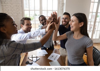 Top view of happy multiethnic young employees join hands give high five celebrating business success at meeting, excited diverse colleagues coworkers involved in teambuilding activity show team spirit