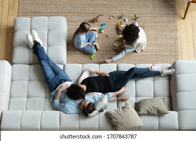 Top view of happy multiethnic young family with small children relax in living room at home together, diverse parents rest on comfortable sofa watch multiracial little daughters play on warm floor