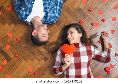 Top view of happy young couple in love lying down on floor and looking at each other. Girl is holding a red paper heart. Valentine's day concept.