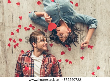 Top view of happy young couple looking at each other and smiling while lying on wooden floor. Girl is holding a red paper heart