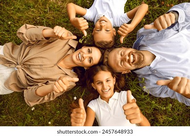 Top view of happy young family of four lying on green grass and showing thumb up sign in summer park. Smiling parents having fun with kids boy and girl in nature enjoying time together. - Powered by Shutterstock