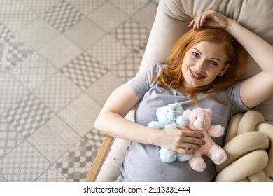 Top View Happy Young Redhead Pregnant Woman Lying On Comfortable Armchair With Two Cute Bear Toys On Tummy. Smiling Female Awaiting Twins Baby Relaxing At Home Dreaming About Happy Motherhood