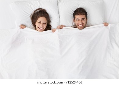 Top view of heads of happy lovely couple lying in bed and smiling broadly after good sex. Lovers enjoying intimacy and togetherness. Just married spending first night at hotel room, feeling excited