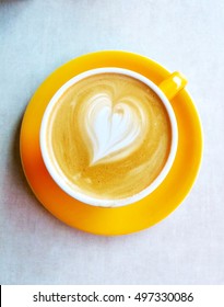 Top View Of A Heart Shaped In A Yellow Cup Of Latte, Cappuccino Coffee. Cafe Art Design With Milk, Cofee, Round Ceramic Glass And Plate For Coffe Lovers - Aka Latte Art.