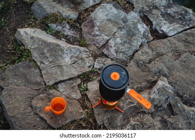 Top View Of Hiking Utensils, Orange Tea Mug Standing On A Stone, Pot For Boiling Water, Set For Kitchen In The Forest