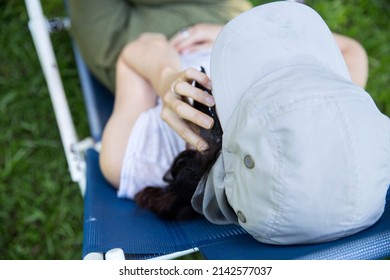 Top View Of Hispanic Young Woman Wearing A Cap Talking With On Mobile Phone And Doing Home Office While Sitting In A Lounge Chair At The Backyard In The Springtime.