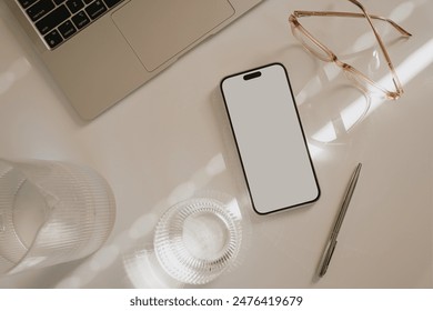 Top view home office workspace. Mobile phone with blank white screen, laptop computer, glasses on white table. Decanter and glass of pure water. Soft sunlight glare and shadow. Mock up with copy space - Powered by Shutterstock