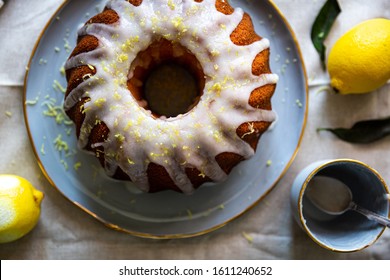 Top view of homemade lemon bundt cake decorated with white icing and lemon zest on pale blue plate with lemons on linen fabric.