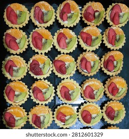 Top view of homemade mini fruit tartlets arranged on a black tray, featuring fresh strawberries, kiwi, and mandarin orange slices on creamy custard filling with golden crispy pastry shells. Colorful dessert concept for bakery, catering, and sweet food photography