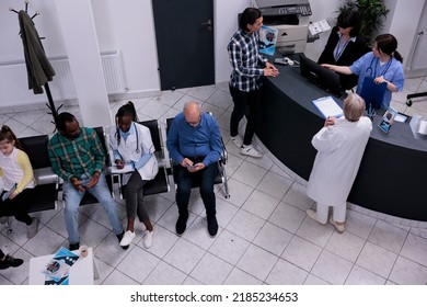 Top View Of Hospital Waiting Area With Senior Doctor Admitting Asian Patient Into Clinic At Front Desk. African American Doctor Filling Form For Patient Appointment In Medical Healthcare Facility.