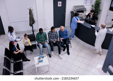 Top View Of Hospital Waiting Room With Diverse Group Of People Talking While Nurse Is Checking Appointment For Mother And Child. Hospital Nurse And Senior Doctor Asking Receptionist About Next