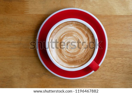 Top view of hot coffee cappuccino latte cup on red ceramic saucer with stirred spiral milk foam on wood table background.