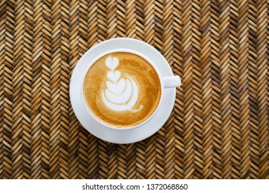Top view of hot coffee latte with heart latte art on rattan table. A cup of cappuccino with beautiful foam in white ceramic cup. Copy space.