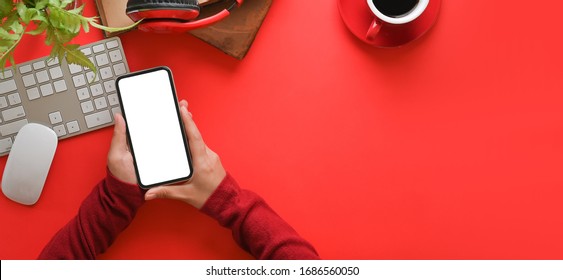 Top view image of hands with red long sleeves holding a empty screen smartphone over orderly working desk. Wireless keyboard, mouse, coffee cup, potted plant, wireless headphone putting on red table. - Powered by Shutterstock