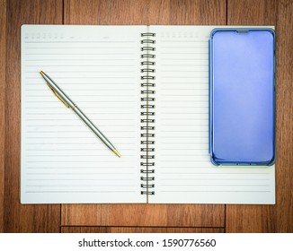 Top view image of open notebook with blank pages and cellphone on wooden table background for adding text or mockup