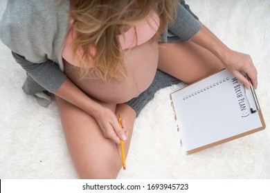 Top view image of pregnant woman holding birth plan checklist and pencil while sitting on her bed at home. Maternity, pregnancy, new life and family planning concept