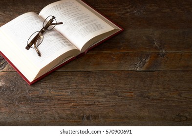 A top view image of a single open book and pair of old eye glasses on a old wooden table top. - Powered by Shutterstock