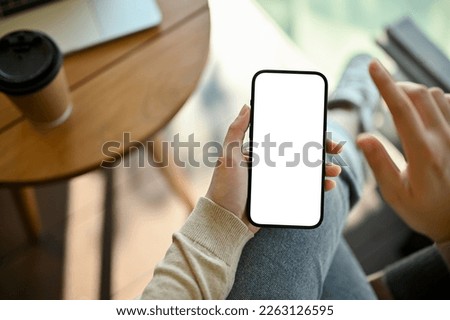 Top view image of a smartphone white screen mockup for display your graphic banner on screen is in a woman's hand. A woman sits in the cafe and uses her phone.