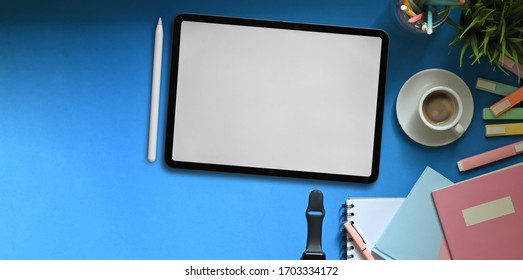 Top View Image Of White Blank Screen Tablet Putting On Colorful Working Desk That Surrounded By Marker Pens, Notebooks, Smartwatch, Coffee Cup, Pencil Holder And Potted Plant. Cluttered Workspace.