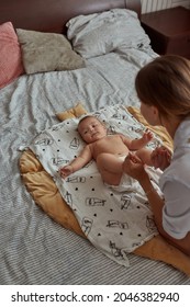 Top View Infant Baby On Parent's Bed During Morning Exercise Routine. Young Woman Holding Baby's Bare Feet In Her Hands. Cute Healthy Baby Looking At Camera.