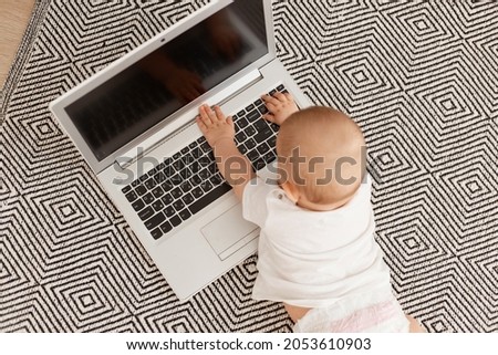 Top view of infant baby wearing white t shirt lying on floor on gray carpet on tummy with laptop with blank screen, small toddler baby using computer.