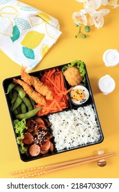 Top View Japanese Bento Lunchbox With Steamed Vegetable, Boiled Egg, Nugget, Sausage, Edamame, And Tempura. On Yellow Background