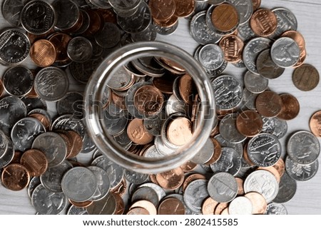 A top view of a jar filled with assorted coins surrounded by scattered change on a wooden surface.