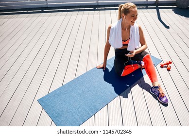 Top view of joyful woman resting during training with dumbbells in open air. She is sitting on mat with towel and holding smartphone while enjoying audio tracks on it with headphones. Copy space in - Powered by Shutterstock