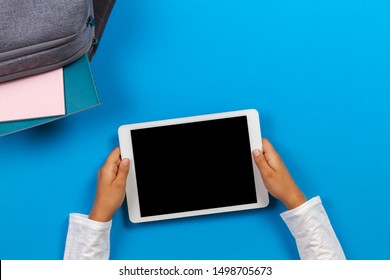 Top view to kid hands with tablet computer, backpack with books and notebook on light blue background