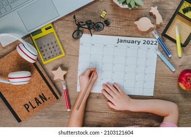 Top View Of Kids Hands Using Calendar For Planning January 2022. Flat Lay Of Workspace. Wooden Table With Laptop, Notebook And Toys. Planning Concept.