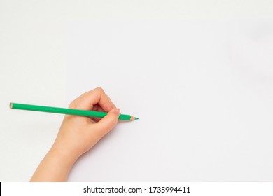 Top View Of Kid's Left Hand Holding Color Pencil And Drawing On White Blank Paper. Copy Space For Text. Mockup.