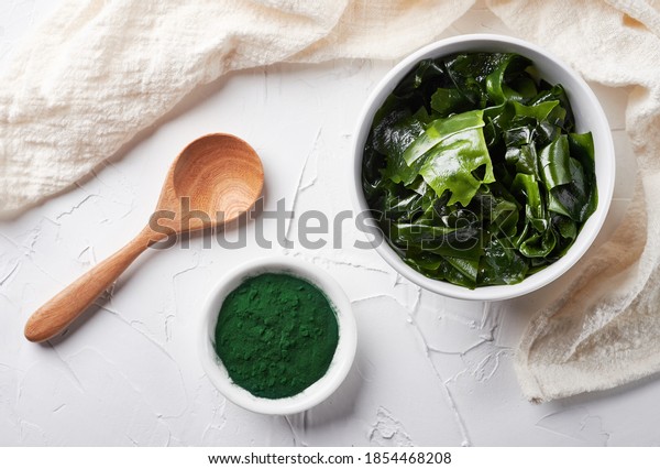 Top view laminaria or kelp\
seaweed and spirulina powder in white bowl and spoon background.\
flat lay spirulina powder with konbu and alga food on white\
background.