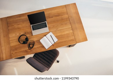 Top View Of Laptop With Blank Screen, Headphones And Notebook On Wooden Table With Office Chair Near By