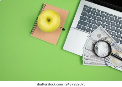 Top view of laptop dollars magnifying glass notepad and apple on green background with empty area for text or marketing. - Powered by Shutterstock